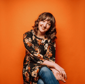 A brunette haired woman smiling and sitting in front of an orange background